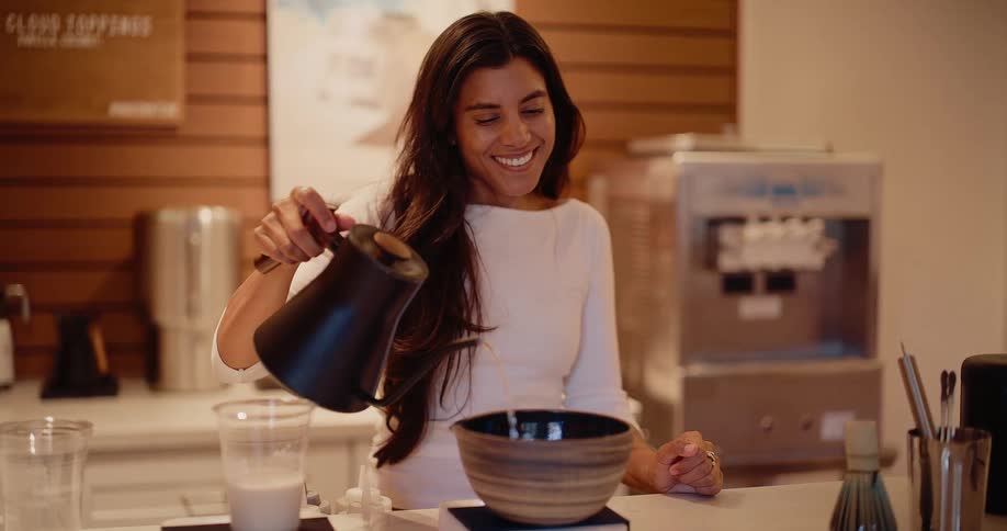 A smiling barista gracefully pours hot water from a gooseneck kettle into a traditional bowl, artfully preparing a matcha tea in slow motion