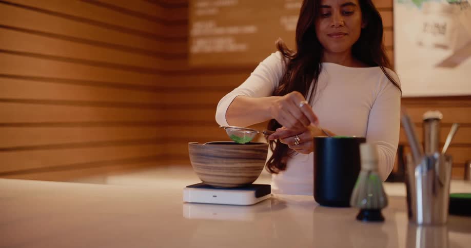 A woman carefully sifts green matcha powder into a wooden bowl in a modern cafe in slow motion