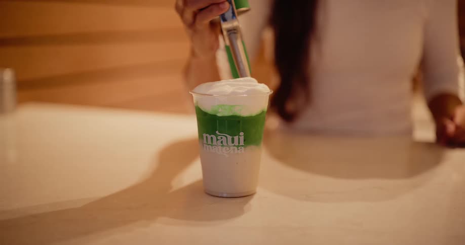 Close-up slow motion of a barista topping a layered green Maui Matcha latte with whipped cream from a dispenser