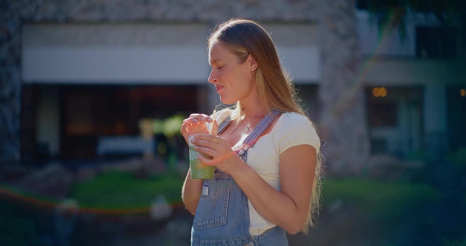 A happy pregnant woman in denim overalls enjoys a healthy green smoothie outdoors on a beautiful sunny day in slow motion