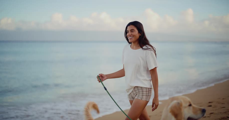 A beautiful young woman smiles while walking her adorable golden retriever on the beach at sunrise in slow motion