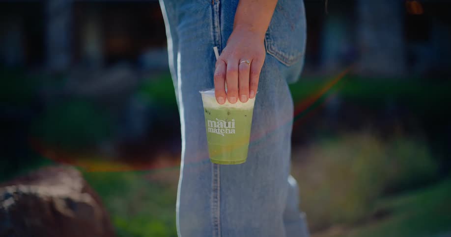 Close up of a woman's hand holding a branded Maui Matcha iced tea while standing outdoors in the sun