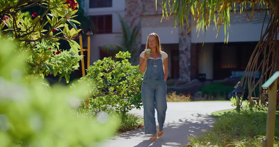 A happy woman in denim overalls enjoys a healthy green smoothie while walking through a lush tropical resort garden