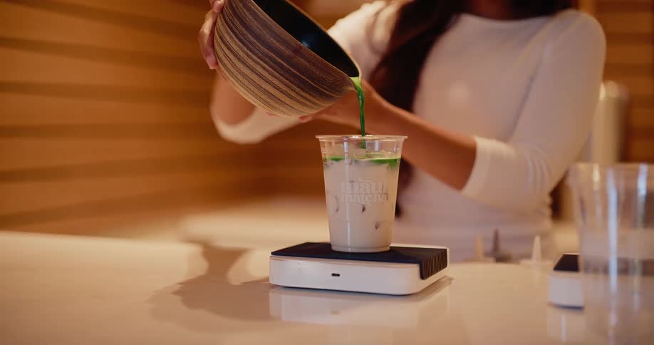 A barista in a modern cafe slowly pours vibrant green matcha into a cup of iced milk