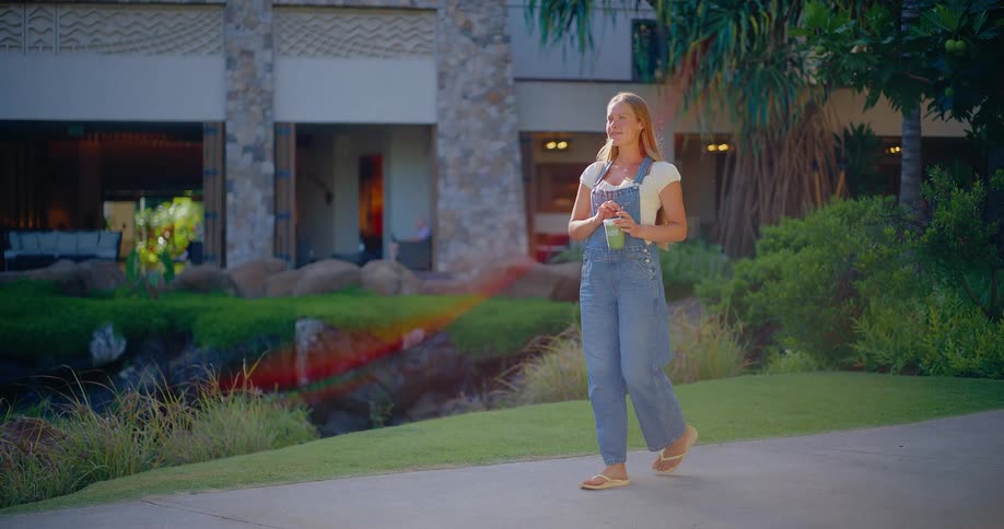 A happy young woman in denim overalls enjoys a healthy green smoothie while taking a leisurely stroll through a lush tropical resort from dusk into a bright sunny morning