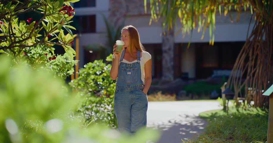A relaxed young woman in denim overalls walks in slow motion through a sunny tropical garden while enjoying a healthy green smoothie
