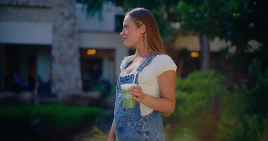 A happy pregnant woman wearing overalls smiles while holding a healthy green smoothie in a sunlit courtyard
