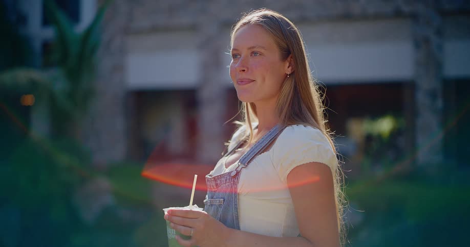 Sun-kissed young woman wearing overalls smiles while holding a healthy green smoothie outdoors on a beautiful day