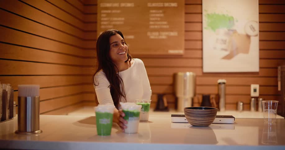 A friendly barista smiles while serving a fresh matcha latte to a pregnant customer in a trendy cafe
