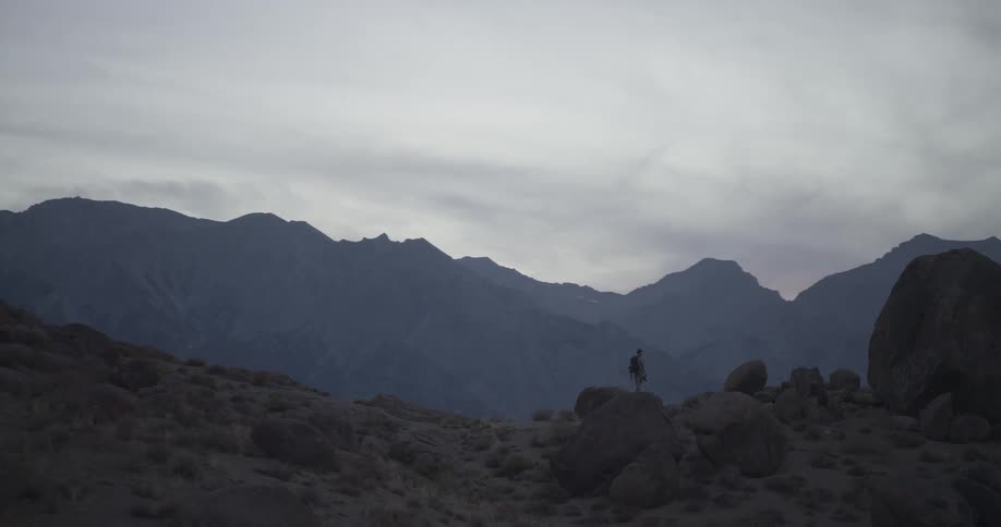 A lone hiker with a backpack stands on a rocky hill, surveying the vast mountain range at dusk