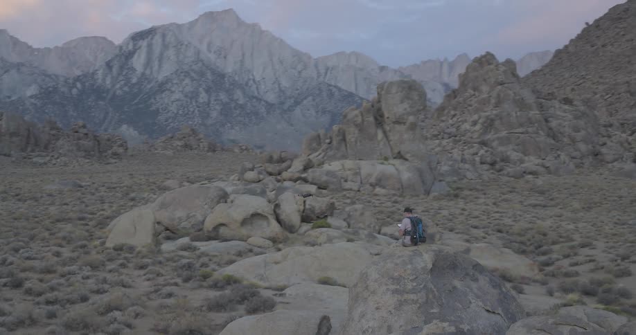 Aerial shot orbits a man with a backpack sitting on a large rock while flying a drone in a vast desert at the base of a mountain range
