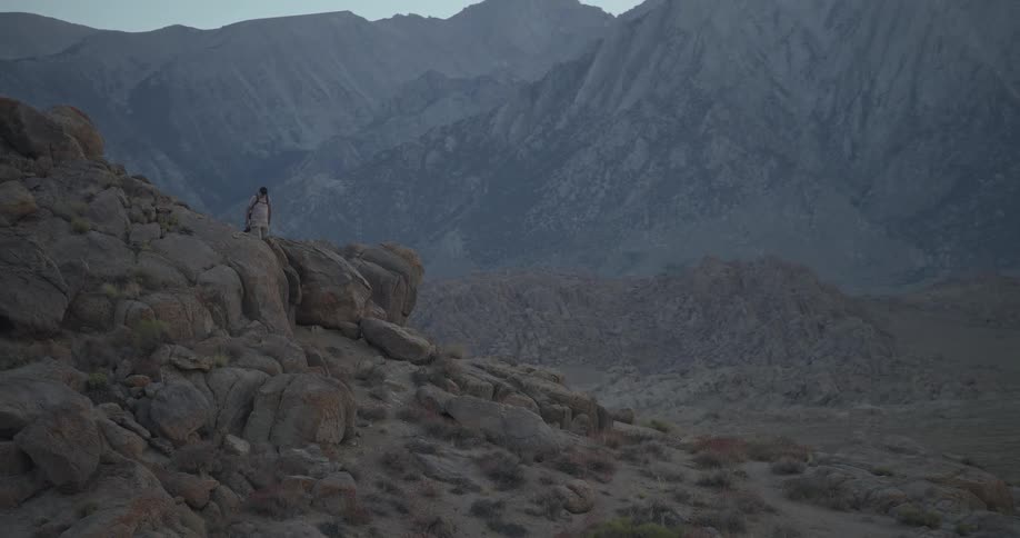 A lone hiker climbs up a rocky mountain outcrop and sits down to enjoy the vast landscape at dusk