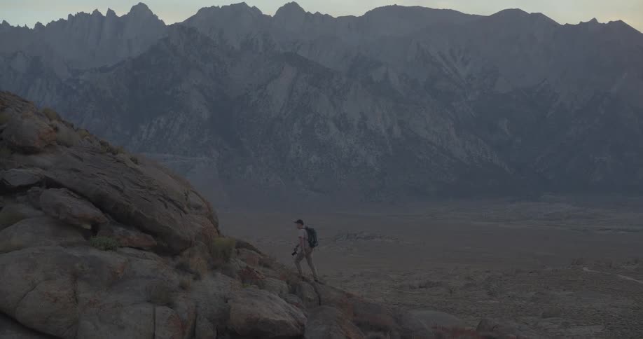 A lone hiker with a backpack carefully navigates a rocky descent in the vast, mountainous wilderness at dawn
