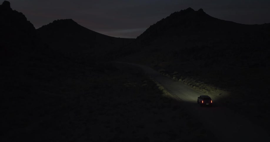 An aerial view of a single car driving along a dark, winding road through a desolate mountain landscape at twilight