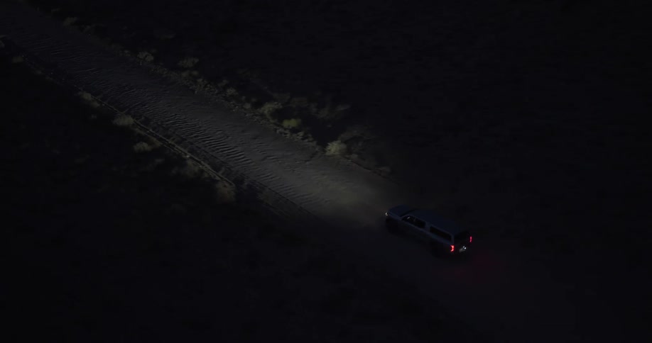 An aerial view shows a lone vehicle driving on a dark, remote dirt road at night, its headlights illuminating the path ahead