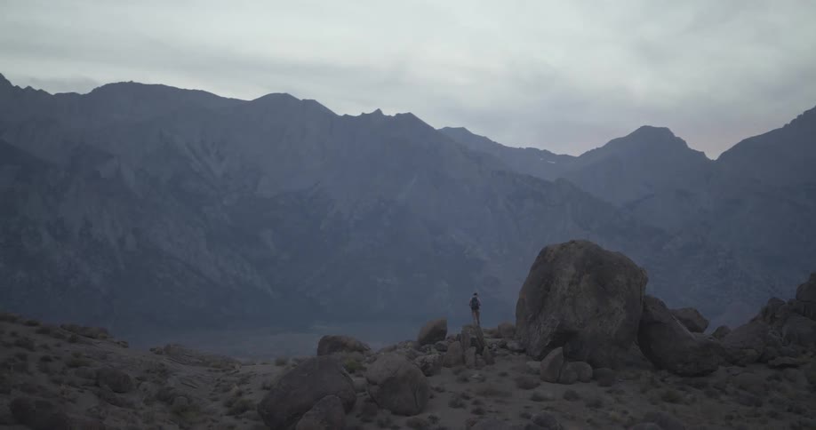 A slow zoom reveals a lone hiker with a backpack standing amidst large boulders, admiring the epic mountain range at dusk