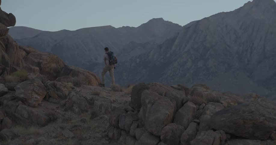 A solo hiker with a backpack traverses a rocky mountain pass at dusk before stopping to photograph the majestic peaks