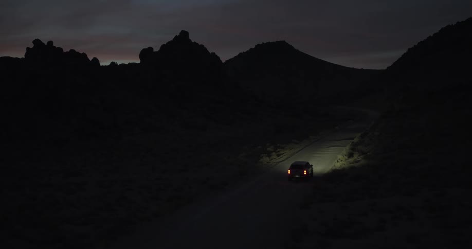 An SUV with its headlights on drives down a remote winding road through a mountain pass at twilight
