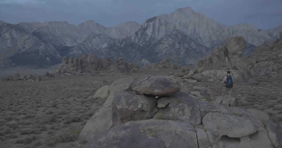 A wide pan shot follows a hiker with a backpack exploring a vast, rocky mountain valley at dusk