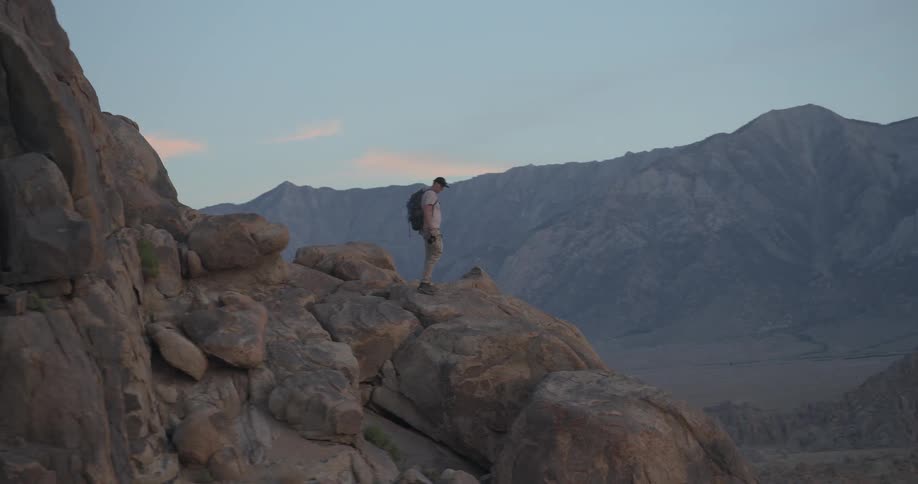 A lone photographer with a backpack hikes along a rocky mountain ridge, stopping to take pictures of the vast landscape at sunset