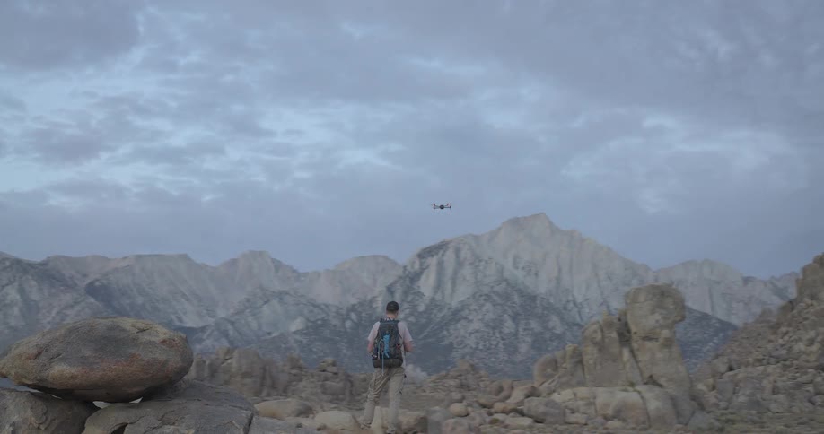 A hiker with a backpack operates a flying drone in a rugged mountain landscape at dusk