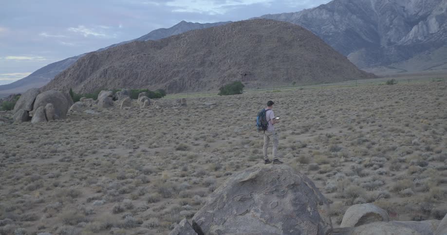 A male adventurer with a backpack stands on a large boulder and flies his drone over a vast rocky desert landscape with mountains in the background