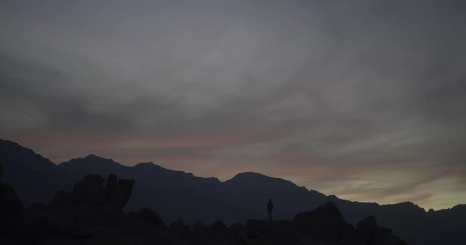 A solitary figure is silhouetted against a dramatic mountain landscape at twilight as the camera pulls back to reveal the epic scenery