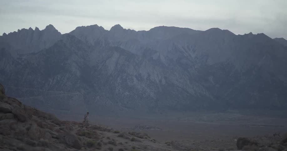 Camera pulls back to reveal a lone hiker walking through a vast desert landscape with a towering mountain range in the background
