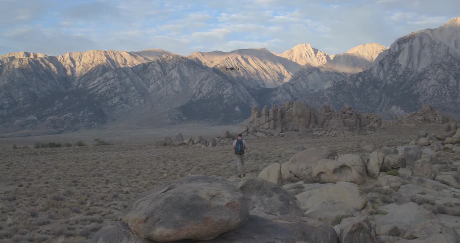 An adventurous hiker stands on a large boulder in the desert and flies a drone to capture the stunning mountain scenery at dawn