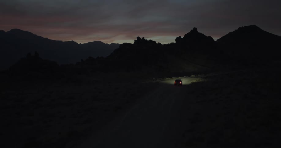 An aerial view follows a lone car with bright headlights driving down a dark, remote desert road toward mountains at twilight