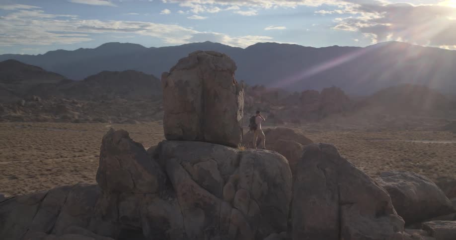 A lone hiker with a backpack climbs onto a large rock formation to enjoy the majestic desert view at sunset
