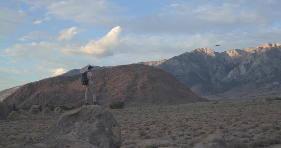 A hiker stands on a large boulder in a vast desert landscape, piloting a drone towards distant sunlit mountains
