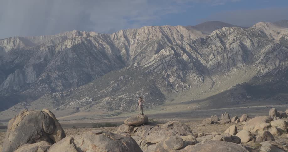 A photographer stands on a large boulder in a vast, rocky landscape to take pictures of the majestic mountains in the distance