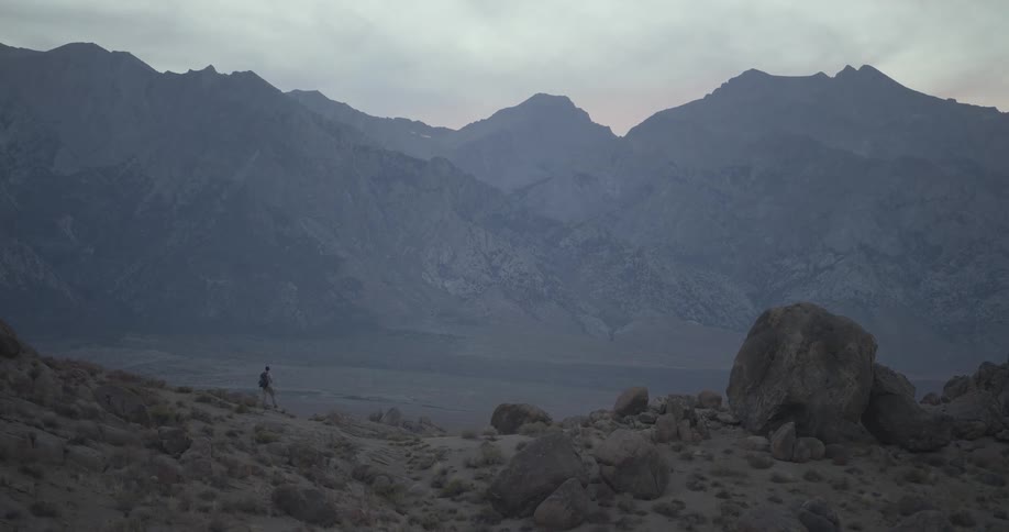 A lone hiker walks across a vast, rocky landscape with a majestic mountain range in the background at dusk