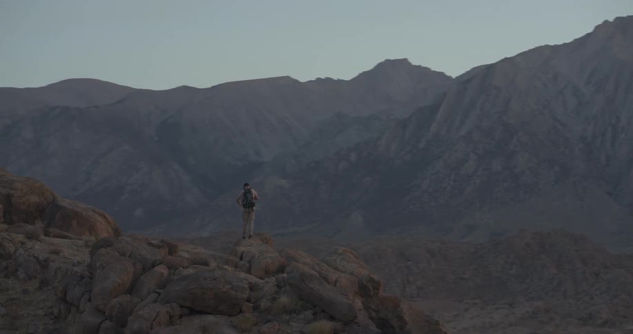 A lone hiker with a backpack stands on a rocky outcrop, admiring the vast mountain range before continuing his trek at dusk