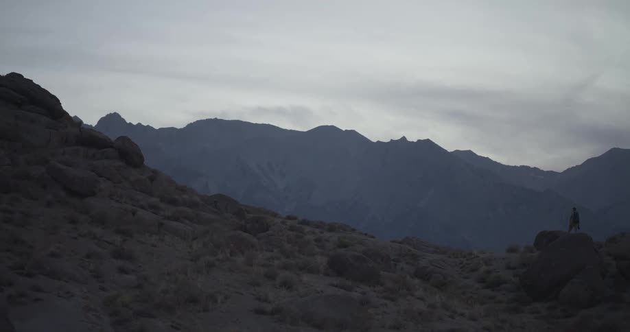 A lone explorer with a backpack hikes across a rocky, desolate landscape with a towering mountain range in the background at dusk