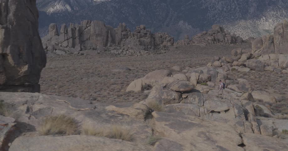 A wide, slow-moving shot captures a lone hiker navigating through a vast and rocky desert landscape