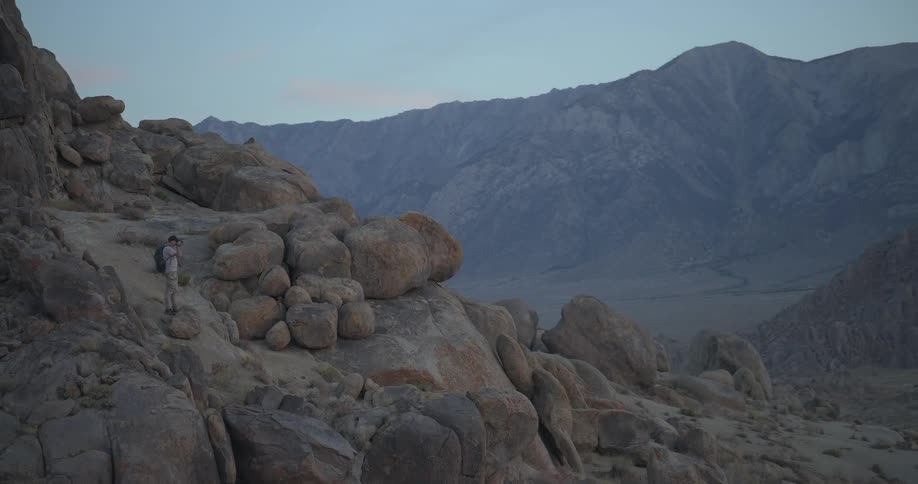 A lone hiker with a backpack and camera navigates a rocky mountain trail at dusk, pausing to photograph the vast landscape