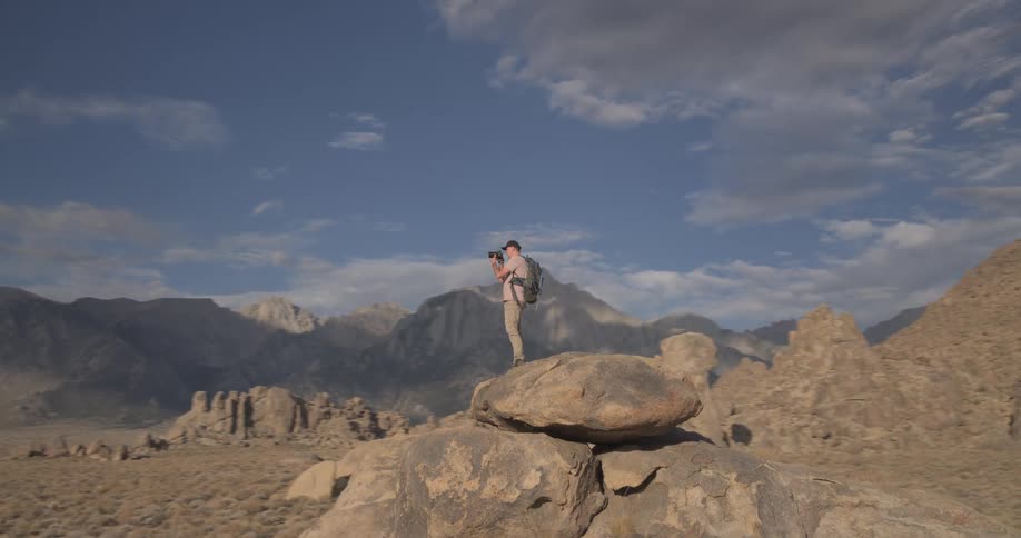 A lone photographer with a backpack stands atop a large boulder, capturing photos of the vast desert and mountain landscape