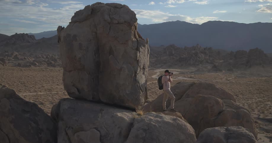 A photographer stands atop a large rock formation in a vast desert valley, capturing the scenic landscape with his camera as the sun shines brightly