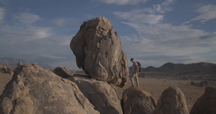 A lone hiker with a backpack explores a vast, rocky desert landscape, carefully navigating around a giant boulder under a cloudy blue sky