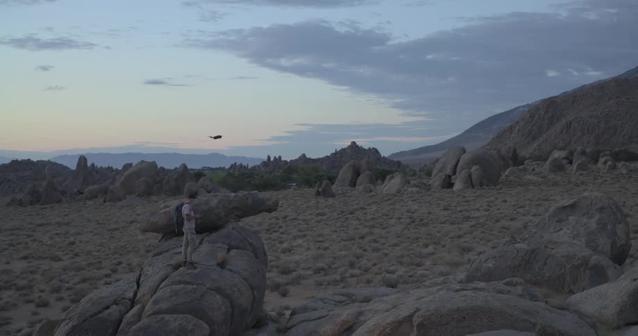 A man wearing a backpack stands on a large boulder and operates a drone in a vast, rocky desert landscape at twilight