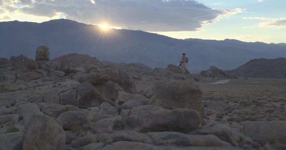 A wide shot of a photographer standing on a large boulder in a rocky desert landscape as the sun sets behind the mountains