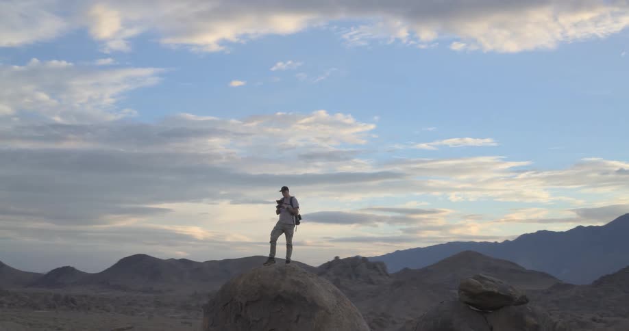 A wide shot pans away from a man standing on a large boulder, operating a drone controller in a vast desert mountain landscape at sunset