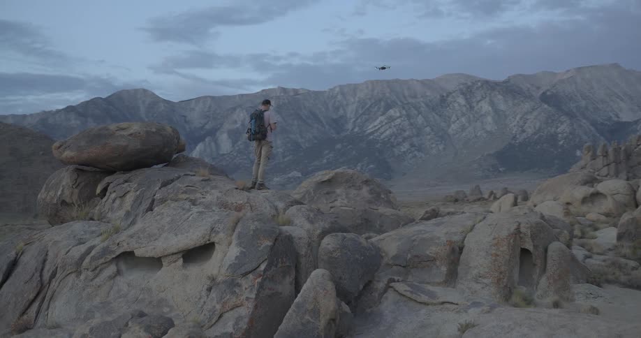 A male hiker with a backpack stands on a rocky precipice and pilots a drone across a vast mountain landscape at twilight