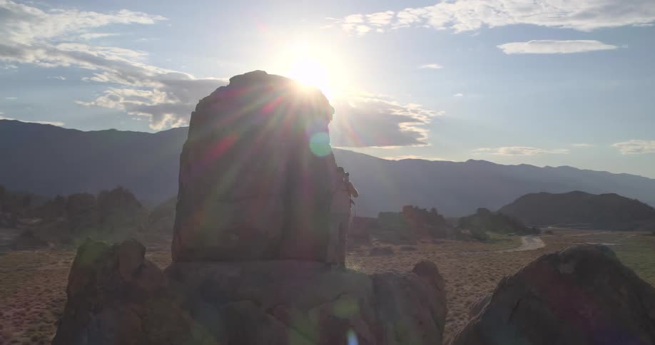 A male photographer climbs a large boulder in the vast desert to take photos of the expansive mountain landscape as the sun sets
