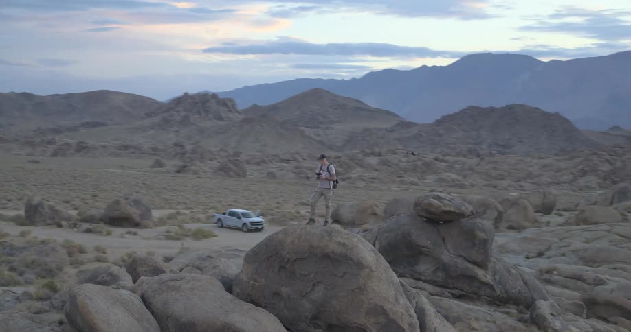 A drone pilot stands on a large boulder in a vast desert landscape, operating his quadcopter at dusk with mountains in the background