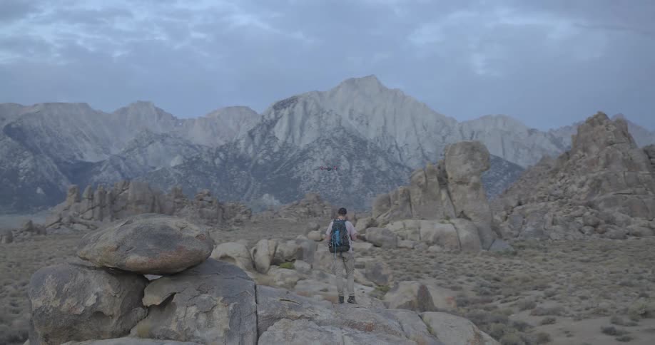 A lone hiker with a backpack operates his drone in a stunningly rugged mountain landscape at twilight