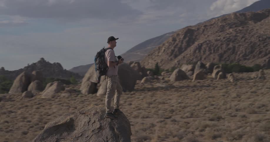 A solo hiker and photographer stands atop a large rock, taking in the vast and rugged mountain landscape as the camera pans around him