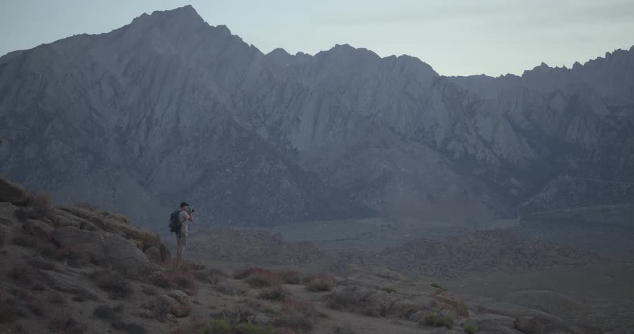 A photographer with a backpack stands on a rocky hill taking photos of a vast mountain landscape at twilight